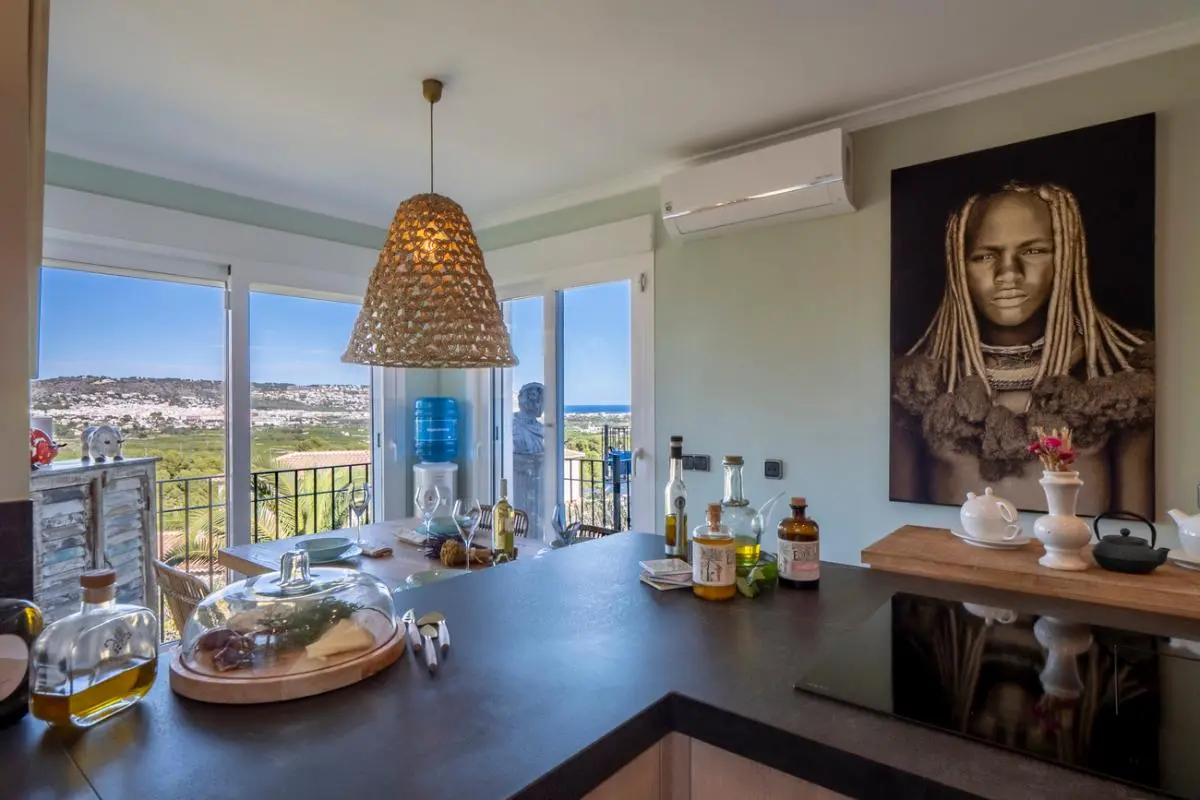 Moderne keuken met donker werkblad, inductiekookplaat en eethoek met een gevlochten hanglamp en panoramisch uitzicht op de kust via glazen deuren.