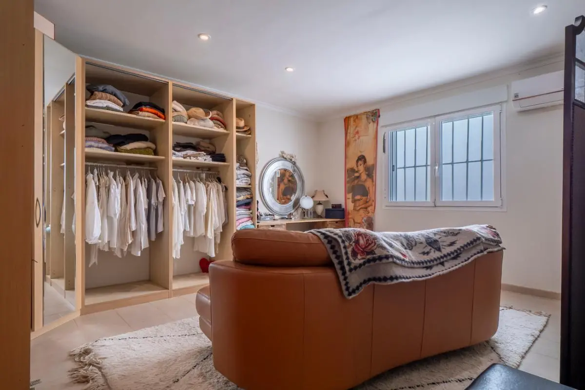 Ruime kleedkamer met een open houten kastsysteem, een cognackleurige leren bank en een kaptafel met ronde spiegel onder inbouwspots.
