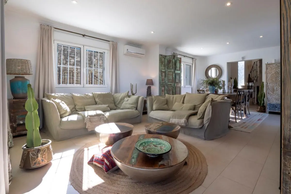 Ruime woonkamer met neutrale banken, ronde houten salontafels op een jute vloerkleed en rustieke decoratie met veel natuurlijk licht.