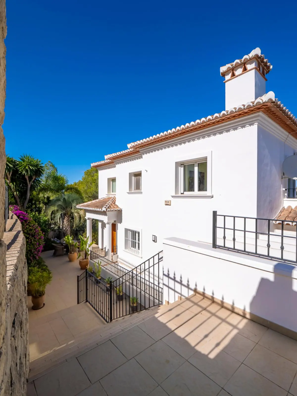Witte mediterrane villa met terracotta dakpannen, een decoratieve schoorsteen en zwart smeedijzeren hekwerk boven een betegeld terras met weelderige beplanting.