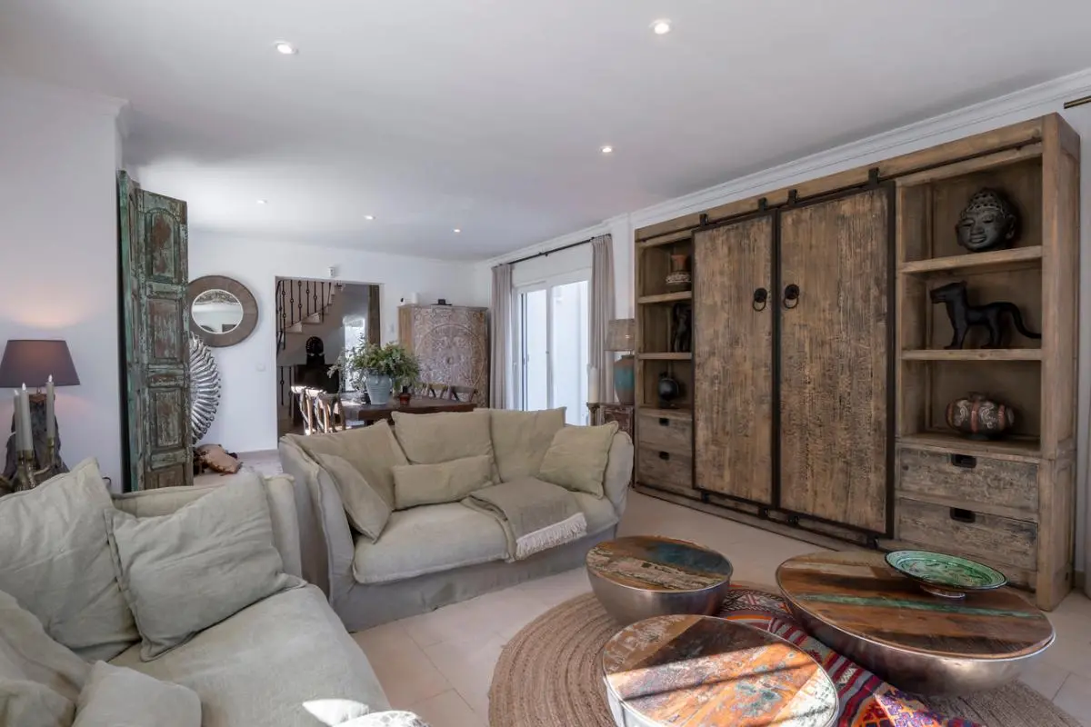 Woonkamer met een grote rustieke houten wandkast, beige linnen banken en ronde salontafels van gerecycled hout op een cirkelvormig jute vloerkleed.