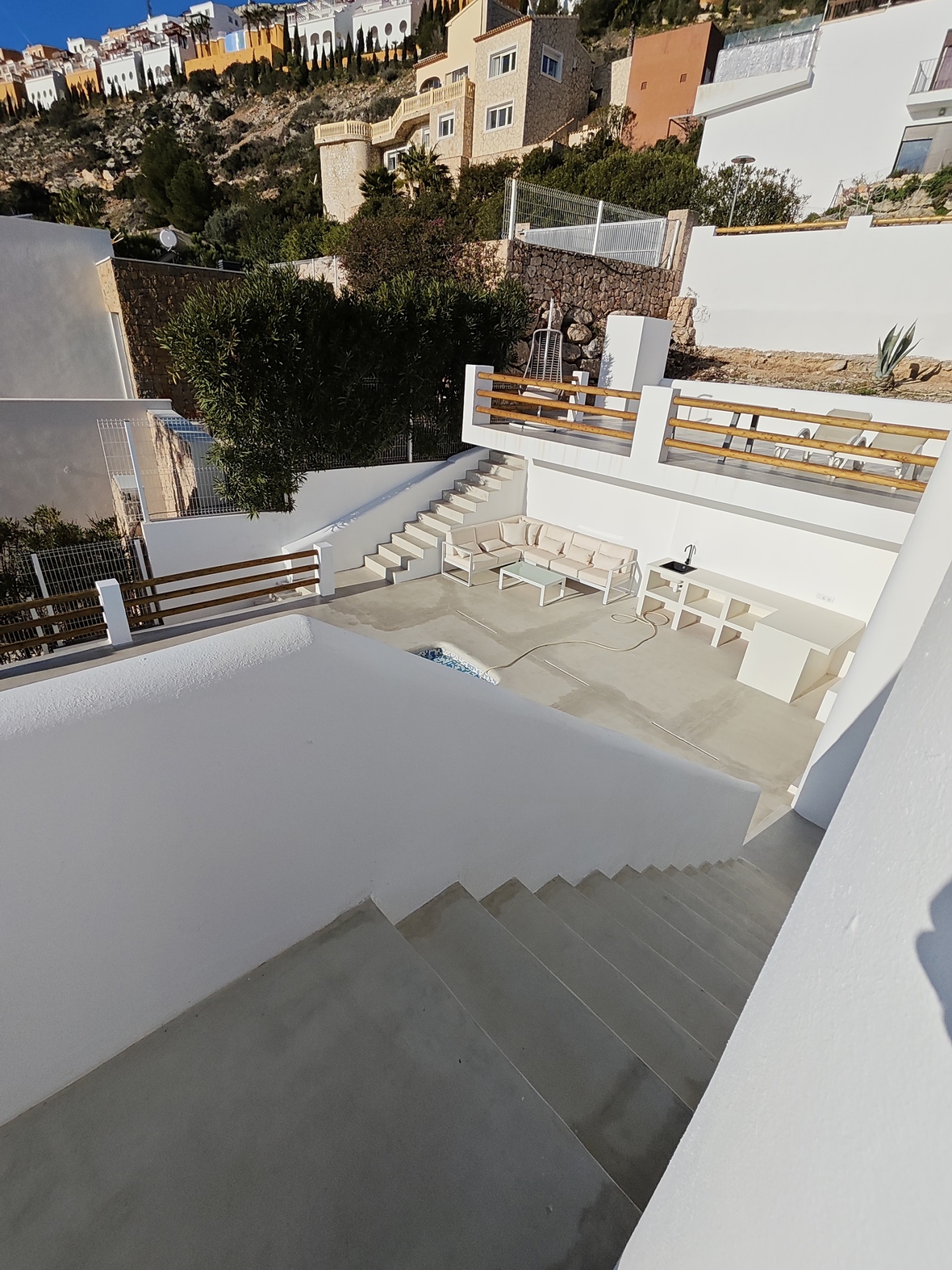 Buitenterras met meerdere niveaus, witte muren, houten leuningen, een zithoek met L-vormige bank en een ingebouwde buitenkeuken tegen een mediterrane heuvel.