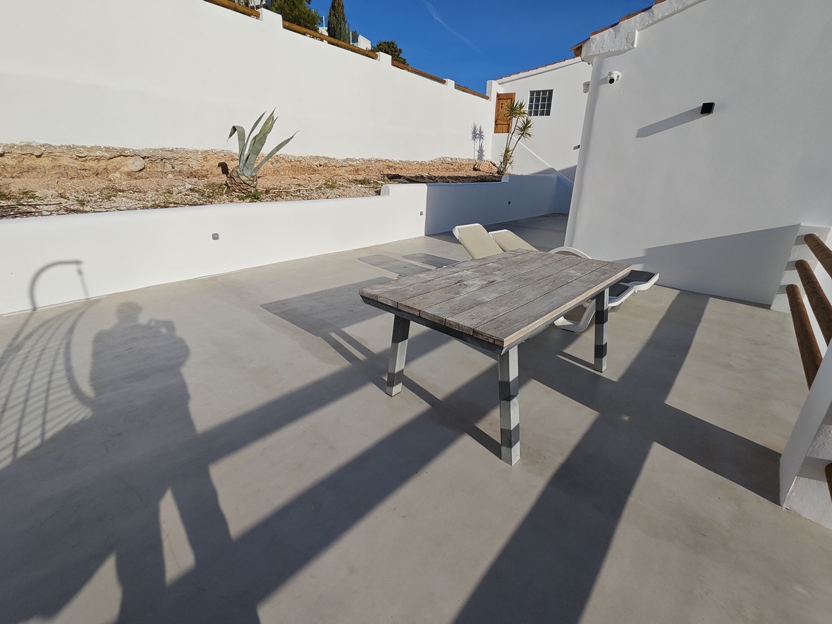 Buitenterras met microcement vloer, voorzien van een verweerde houten tafel en witte muren onder een strakblauwe lucht.