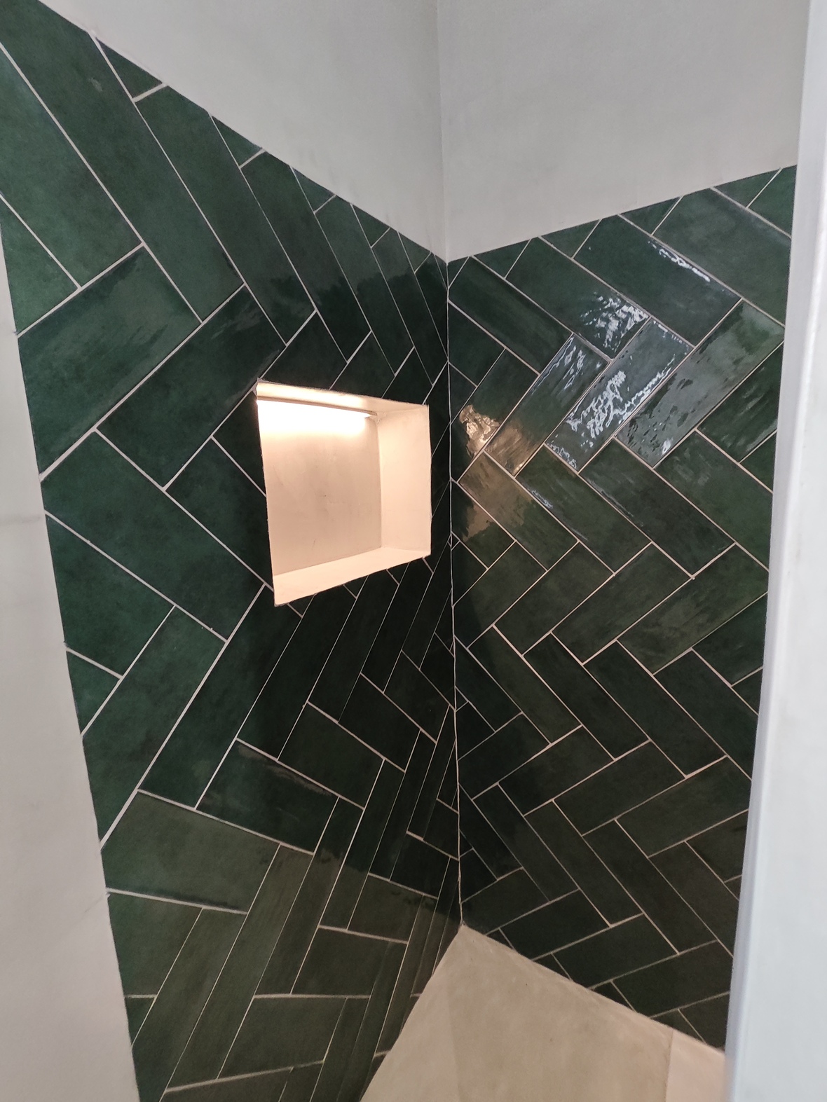 Detail van een douchehoek met glanzende smaragdgroene tegels in visgraatmotief en een verzonken nis met geïntegreerde warme verlichting.