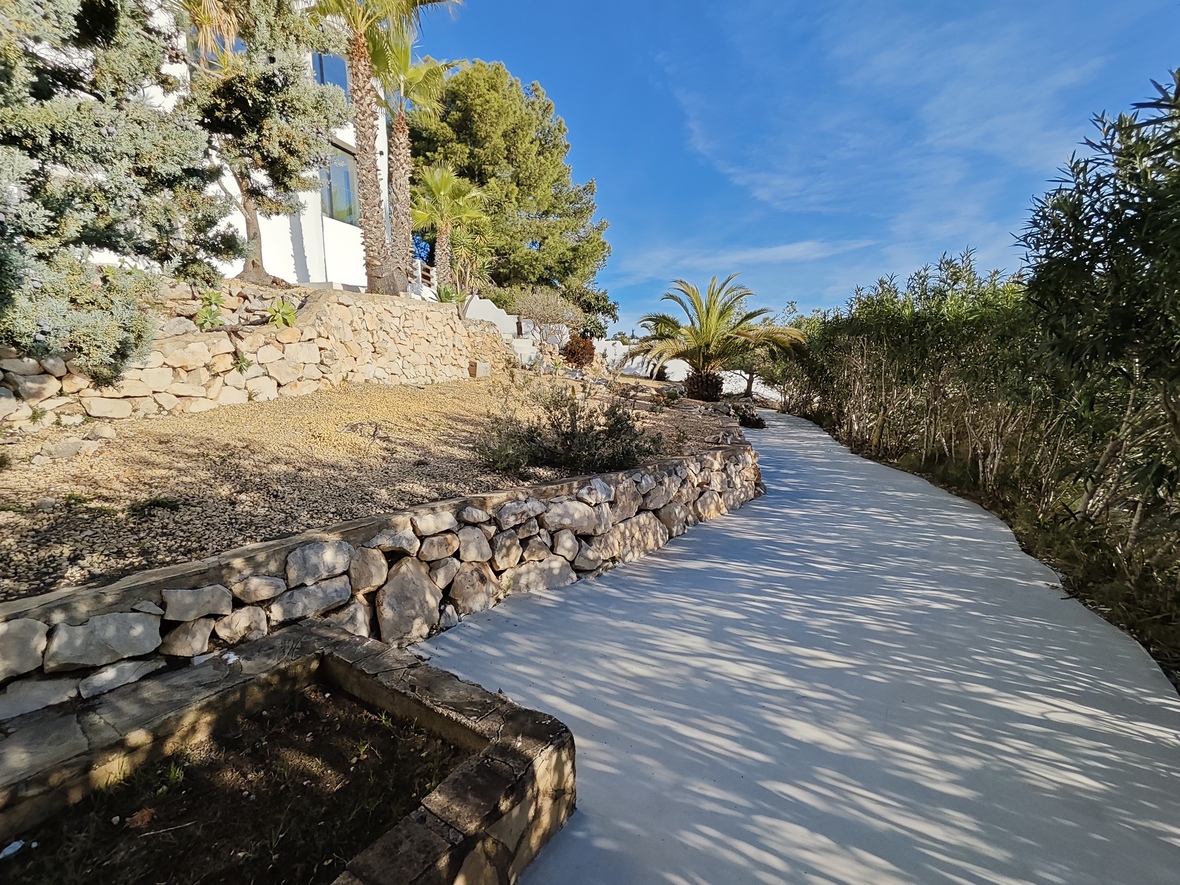 Een glooiend geplaveid pad door een mediterrane tuin met drooggestapelde stenen muren, palmbomen en pijnbomen naast de witte gevel.