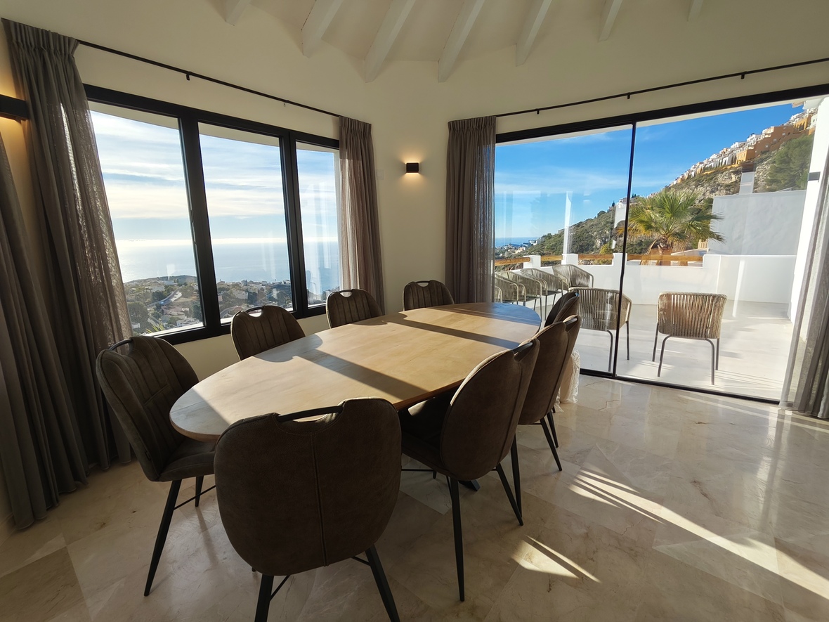 Eethoek met ovale houten tafel en marmeren vloer, voorzien van grote raampartijen met panoramisch zeezicht en directe toegang tot het zonnige terras.