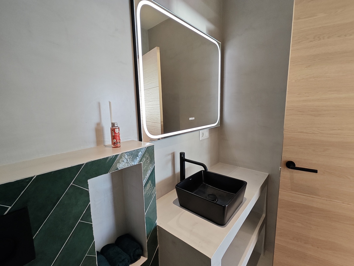 Eigentijdse badkamer met een zwarte opzetwastafel, LED-verlichte spiegel en smaragdgroene visgraattegels op een microcement wand.