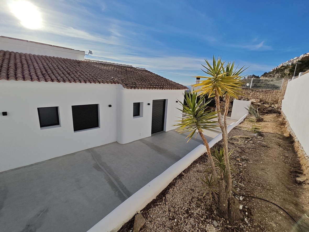 Exterieur van een minimalistische witte villa met pannendak, donkere rolluiken en een betonnen terras grenzend aan een yucca en natuurstenen muur.