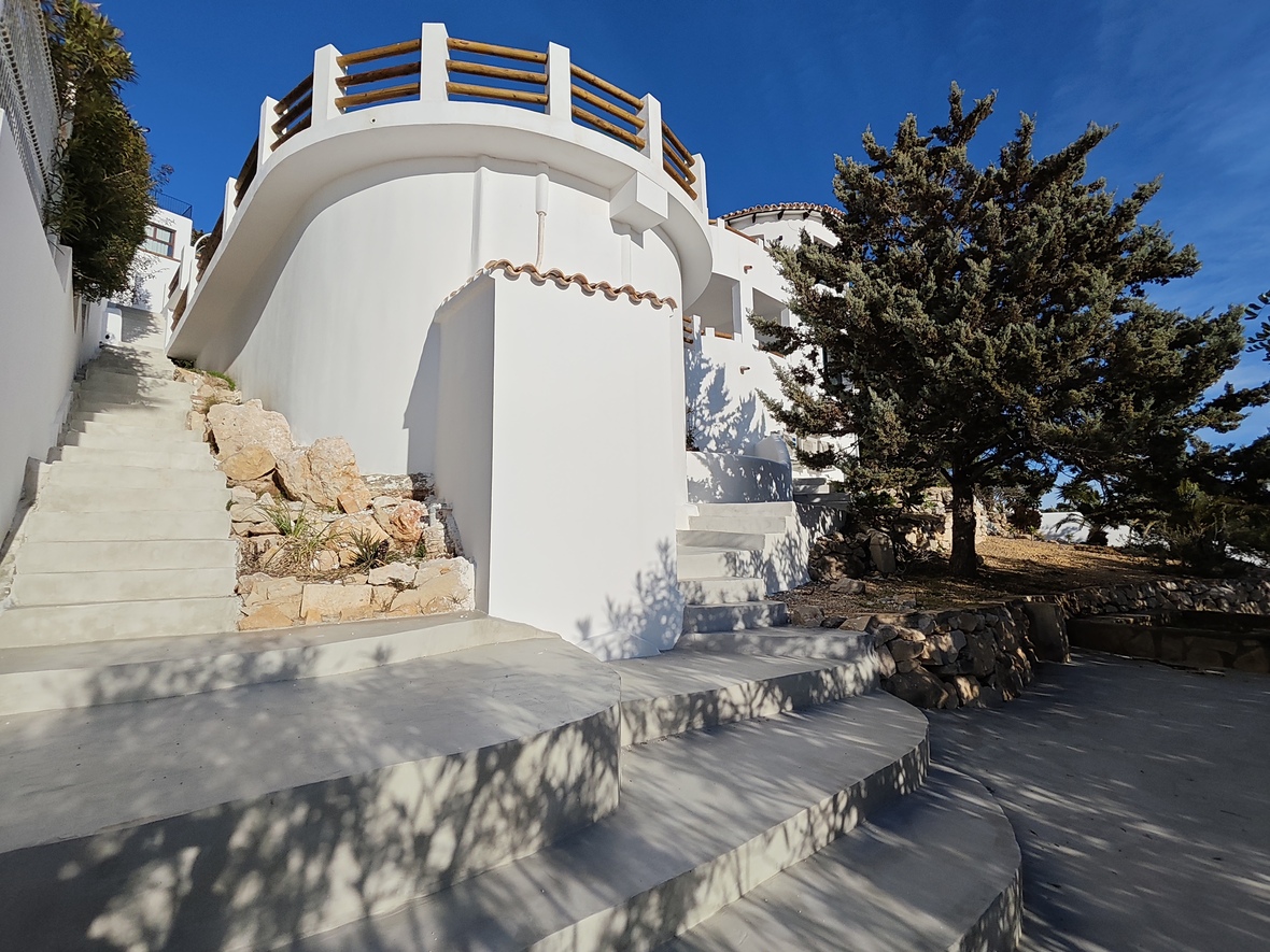 Exterieur van een witte mediterrane villa met gebogen muren, brede betonnen trappen en een rond terras met houten balustrade.