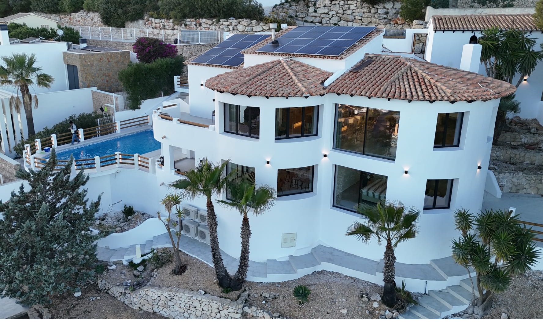 Exterieur van een witte villa met meerdere niveaus, gebogen architectuur, terracotta daken met zonnepanelen en een privézwembad met palmbomen.