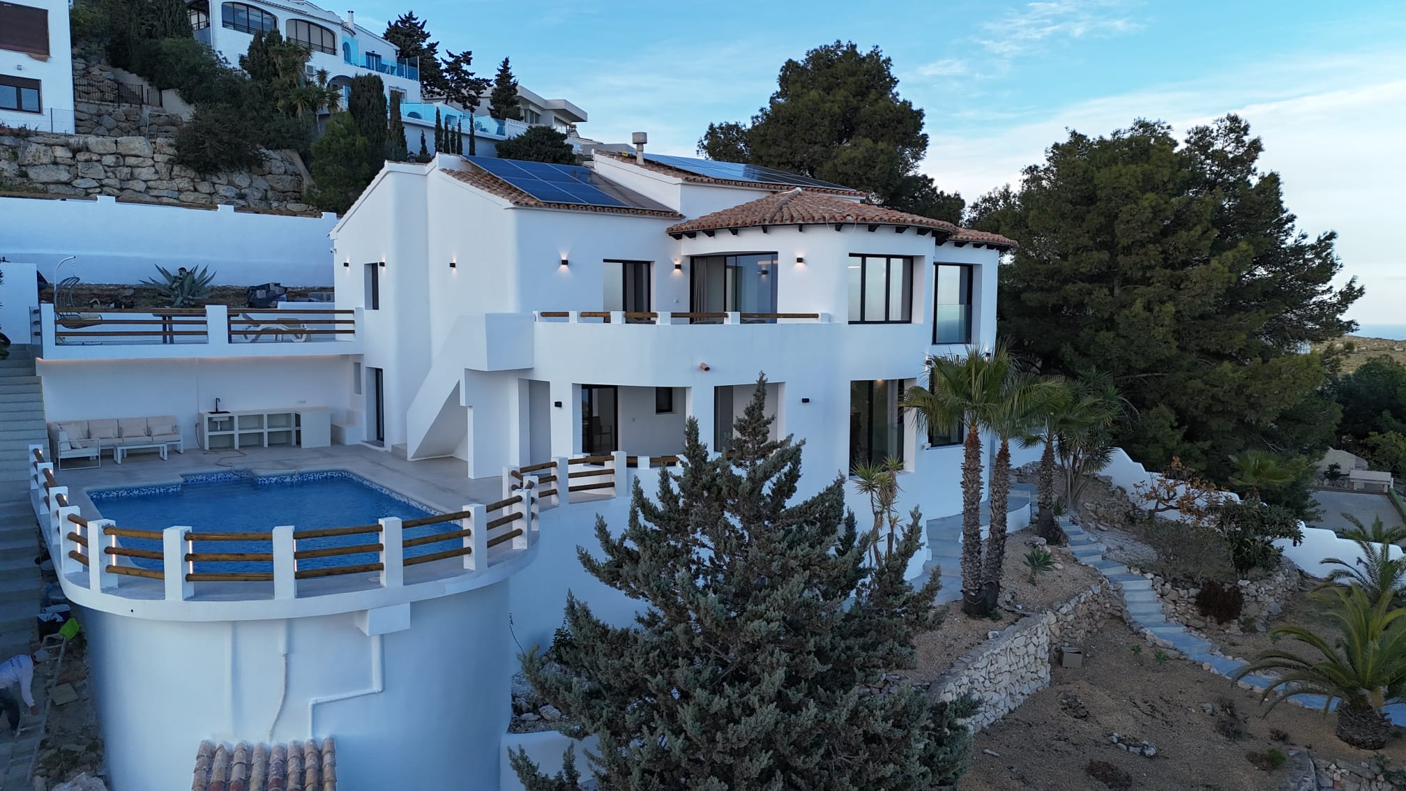 Exterieur van een witte villa met meerdere verdiepingen, een rond zwembad, zonnepanelen op het dak en terrastuinen met mediterrane flora.