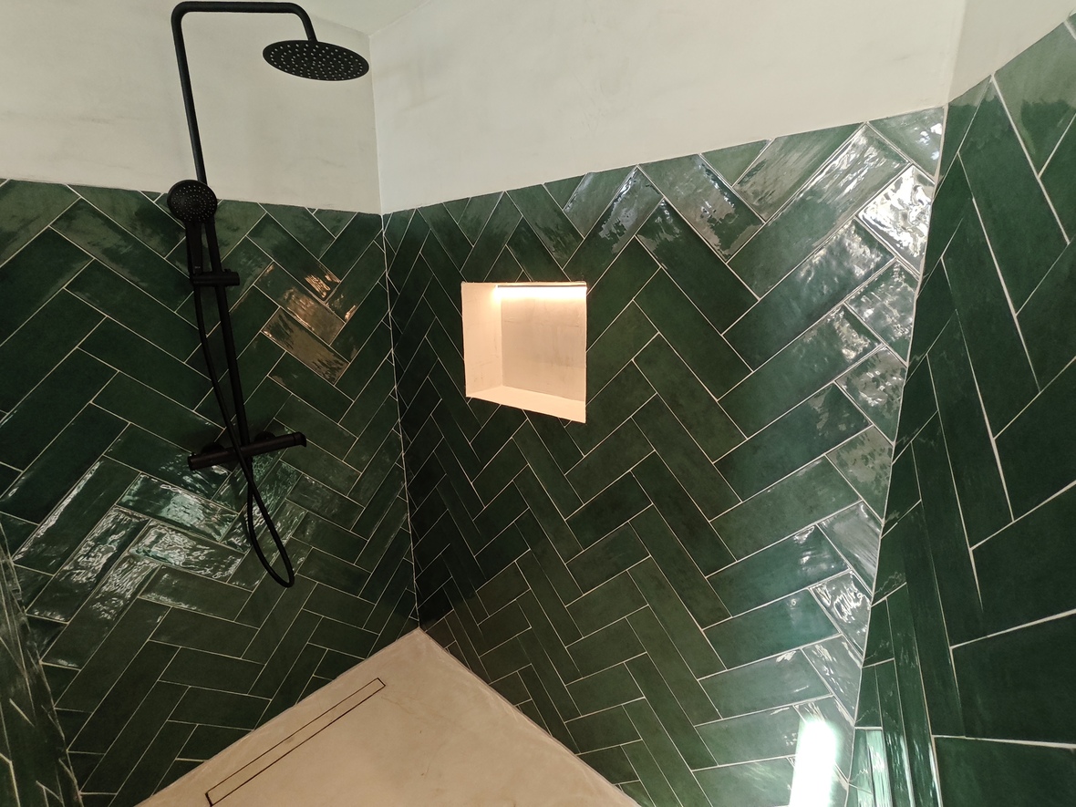 Inloopdouche met smaragdgroene visgraat tegels, matzwarte kranen en een verlichte nis. Voorzien van een lichte vloer met een stijlvolle lijndrain.