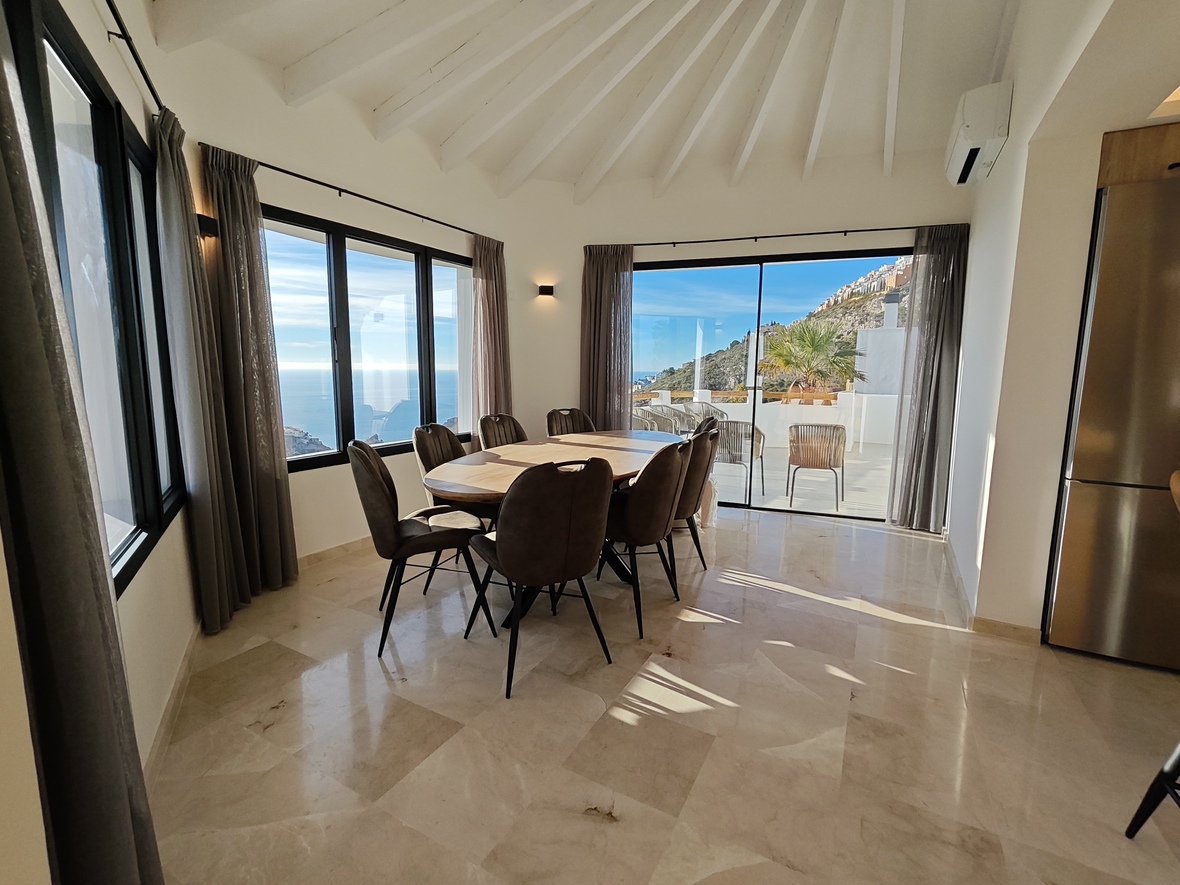 Lichte eetkamer met ovale houten tafel, marmeren vloer en een gewelfd balkenplafond. Grote ramen bieden panoramisch zeezicht en toegang tot het terras.
