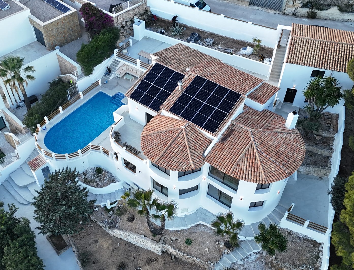 Luchtfoto van een mediterrane villa met terracotta daken voorzien van zonnepanelen, een ovaal zwembad en stenen terrassen verdeeld over meerdere niveaus.