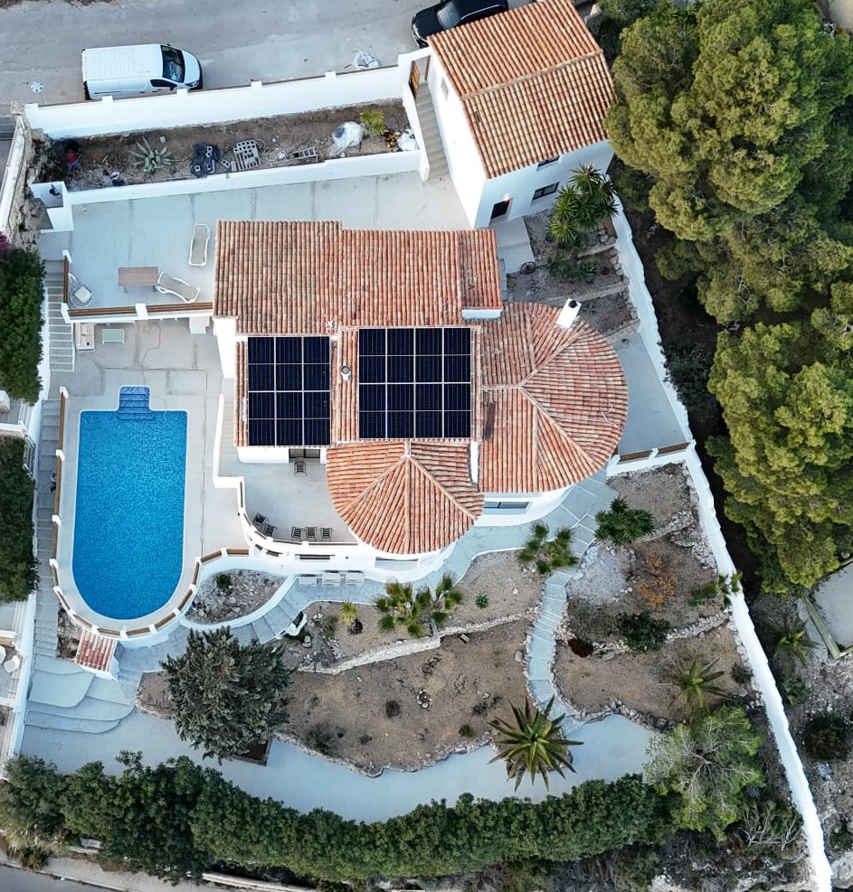 Luchtfoto van een mediterrane villa met zonnepanelen op het pannendak, een gebogen zwembad en terrasvormige tuinen met inheemse beplanting.
