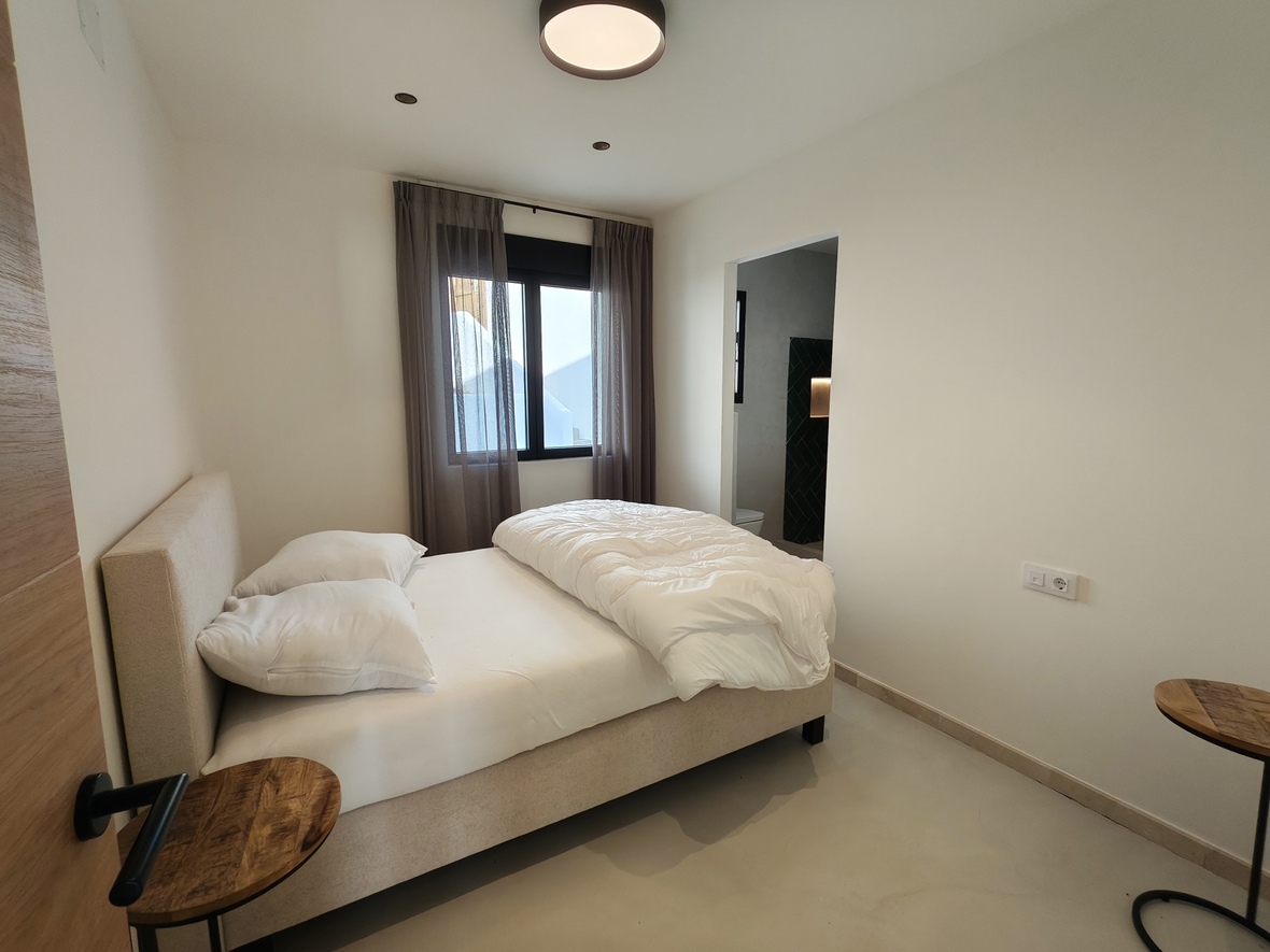 Minimalistische slaapkamer met beige gestoffeerd bed, houten bijzettafels en aangrenzende badkamer met donkere tegels. Voorzien van een gietvloer en zwarte raamkozijnen.