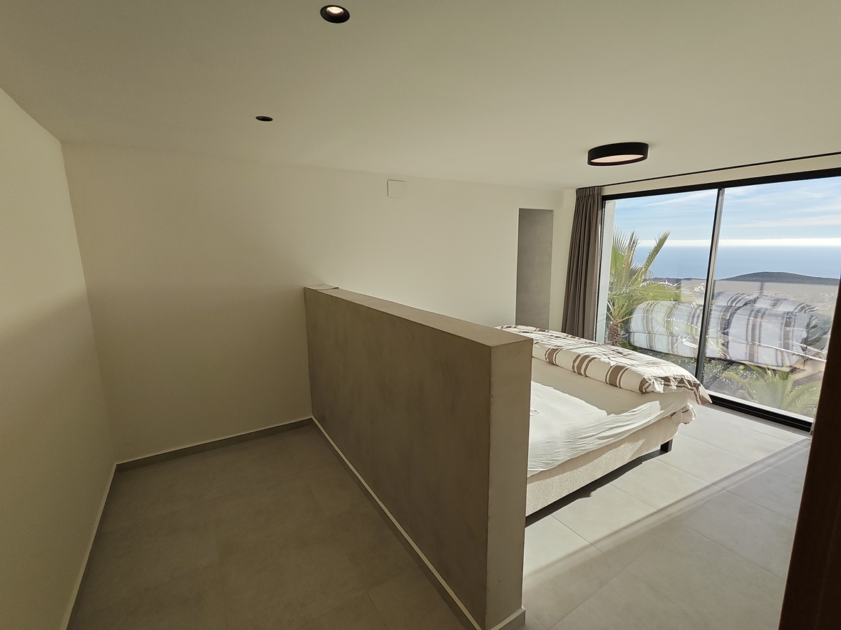 Minimalistische slaapkamer met een lage scheidingswand, grote glazen schuifdeuren met uitzicht op zee en verzonken plafondverlichting.