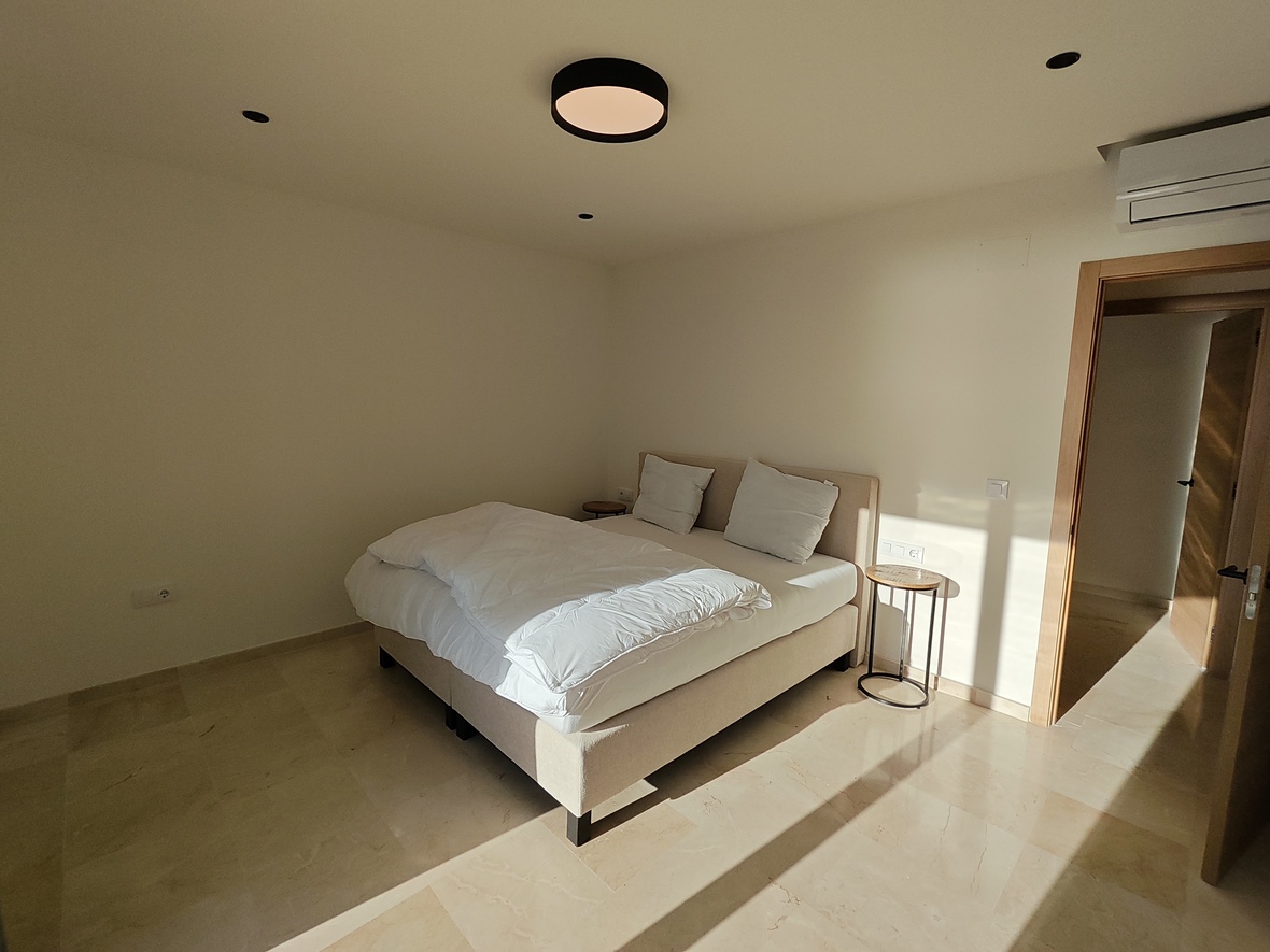 Minimalistische slaapkamer met gepolijste stenenvloer, een gestoffeerd tweepersoonsbed en moderne verlichting met een ronde plafondlamp en inbouwspots.