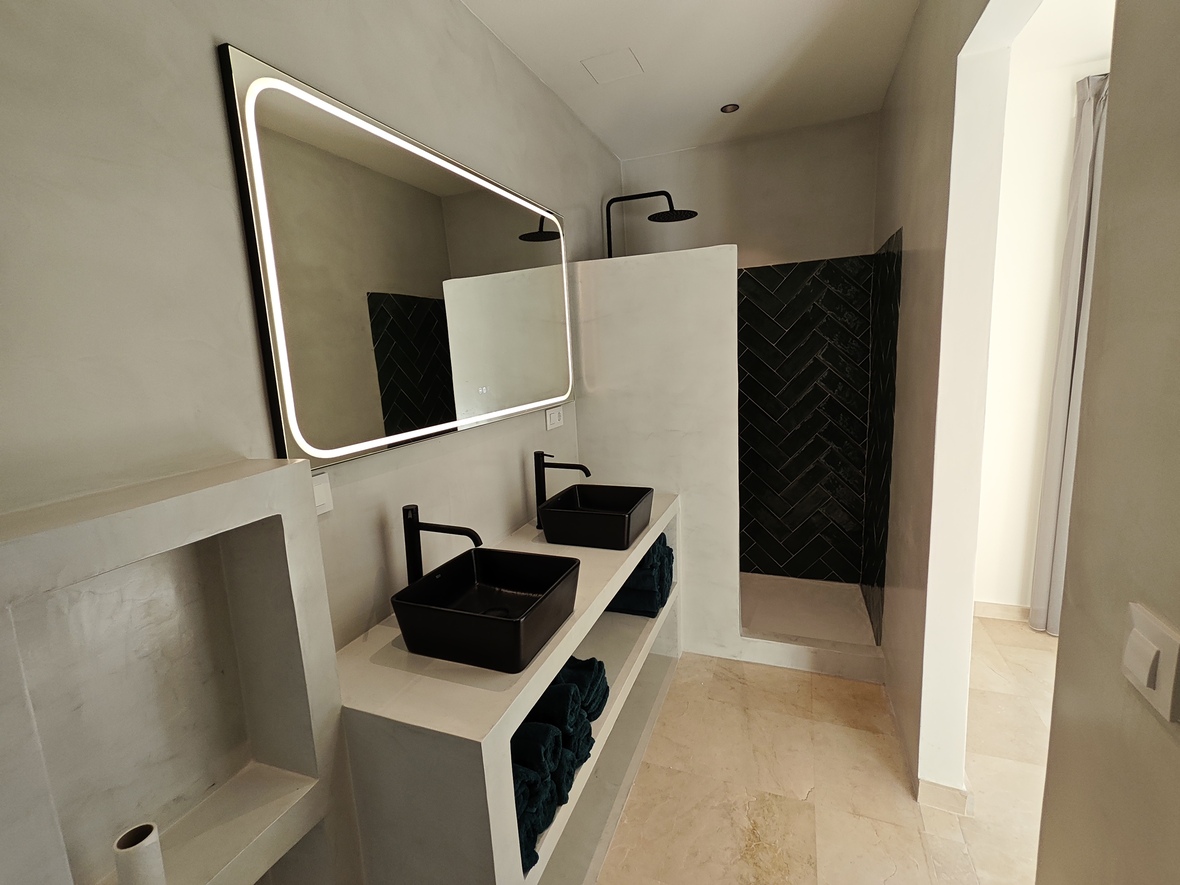 Moderne badkamer met dubbele wastafel van betonciré, zwarte opzetkommen, een verlichte spiegel en een inloopdouche met zwarte visgraat tegels.