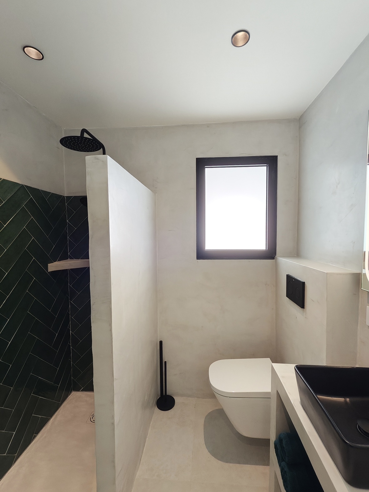 Moderne badkamer met inloopdouche voorzien van groene visgraattegels, hangend toilet en witte microcement wanden met zwarte accenten.