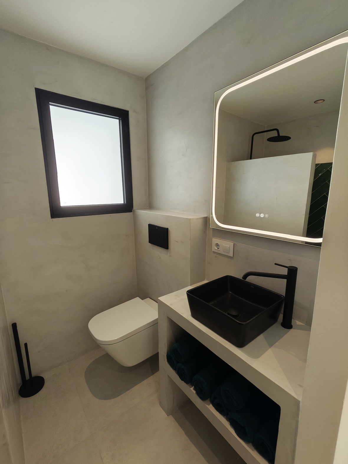 Moderne badkamer met microcement afwerking, een matzwarte opzetwastafel en een verlichte spiegel. Voorzien van zwart deurbeslag en een hangend toilet.