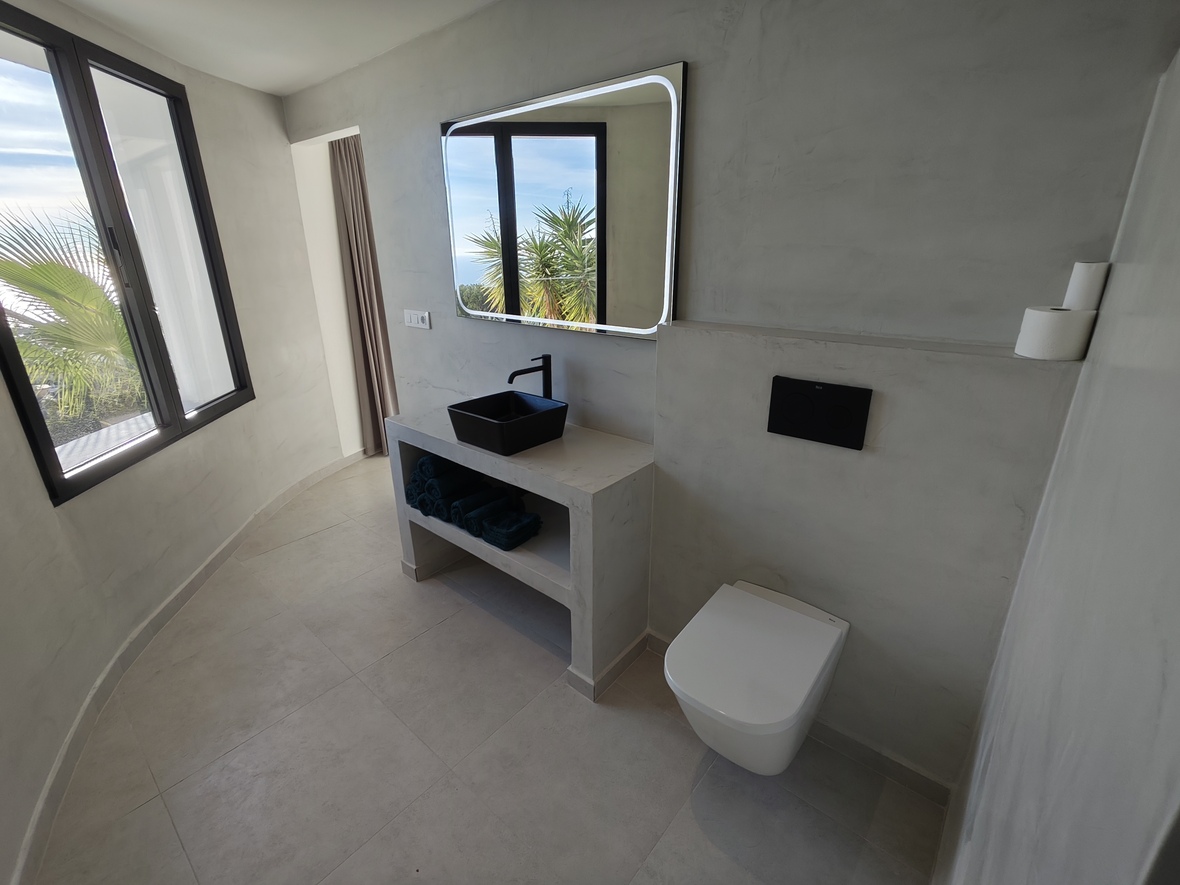 Moderne badkamer met wanden van microcement, een zwarte opzetwastafel en een verlichte spiegel. Grote ramen bieden natuurlijk licht en uitzicht op palmbomen.