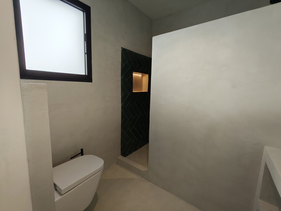 Moderne badkamer met wanden van microcement, hangend toilet en inloopdouche met donkergroene visgraattegels en een verlichte nis.