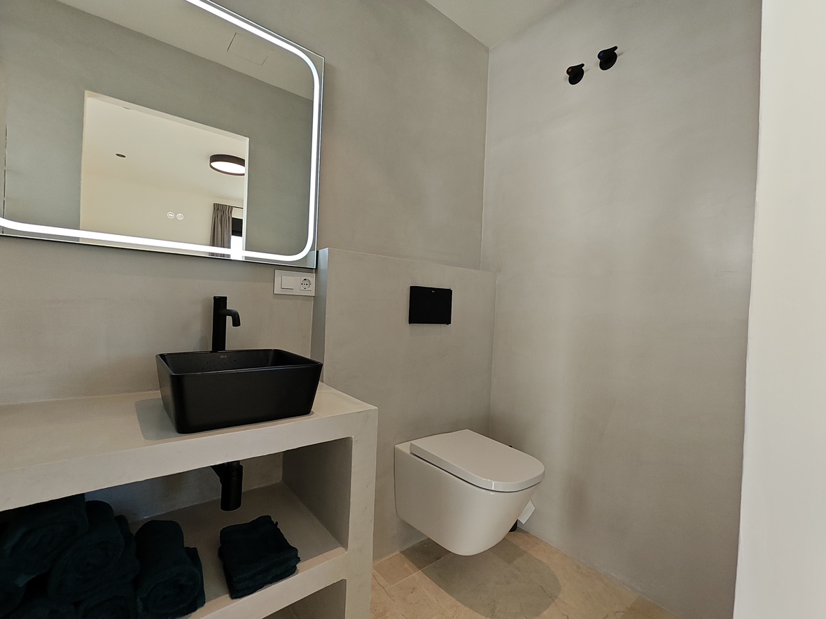 Moderne badkamer voorzien van een zwarte opzetwastafel, verlichte spiegel en hangend toilet tegen wanden van grijs microcement.