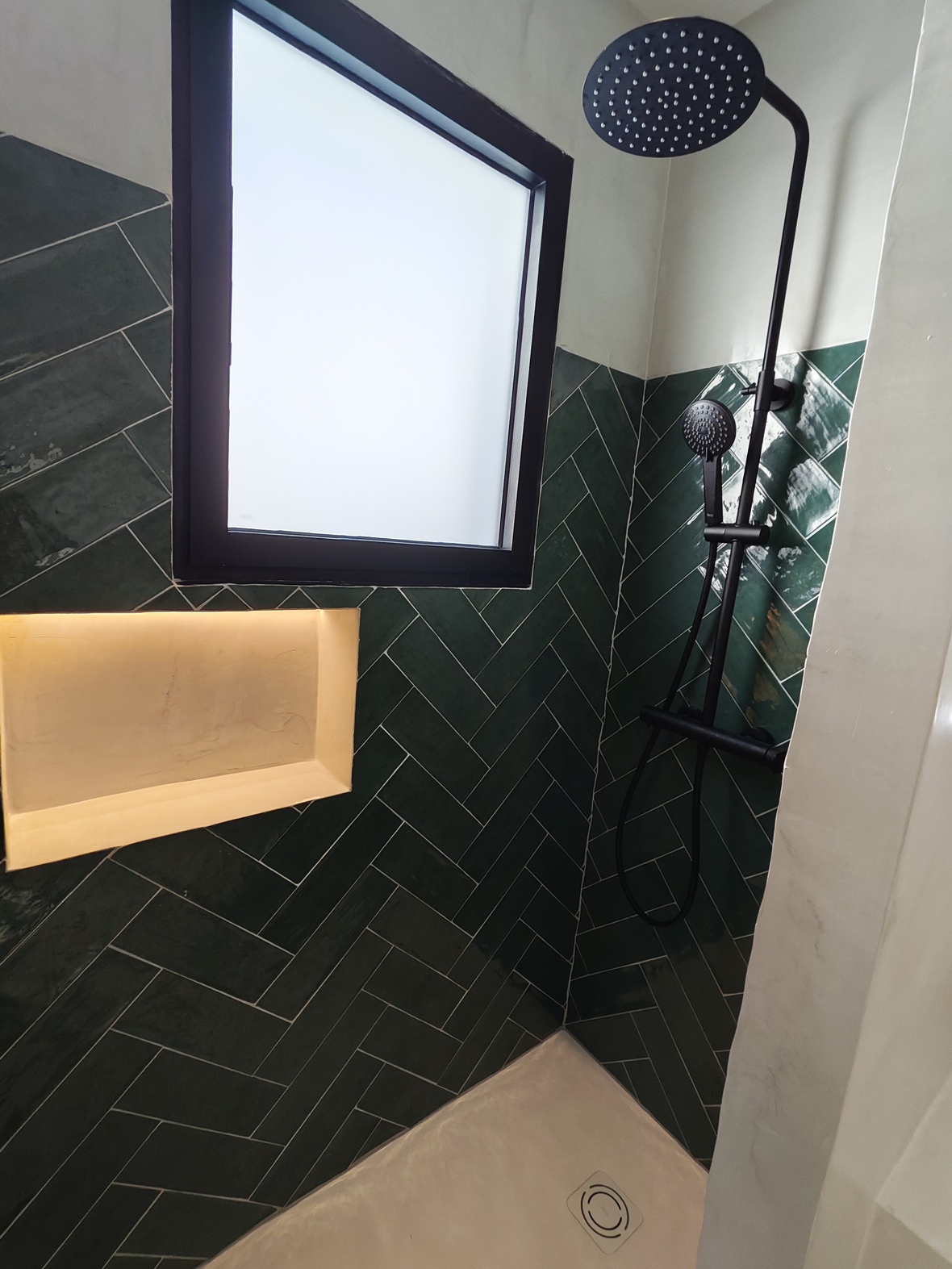 Moderne douche met donkergroene tegels in visgraatmotief, matzwarte kranen, een verlichte nis en een raam met melkglas voor natuurlijk licht en privacy.