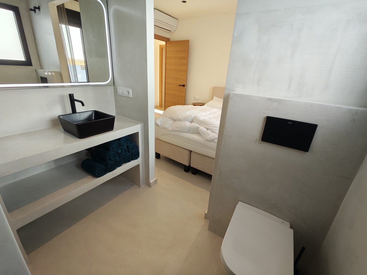 Moderne ensuite badkamer met microcement afwerking, een zwarte opzetwastafel en achterverlichte spiegel, grenzend aan de slaapkamer.