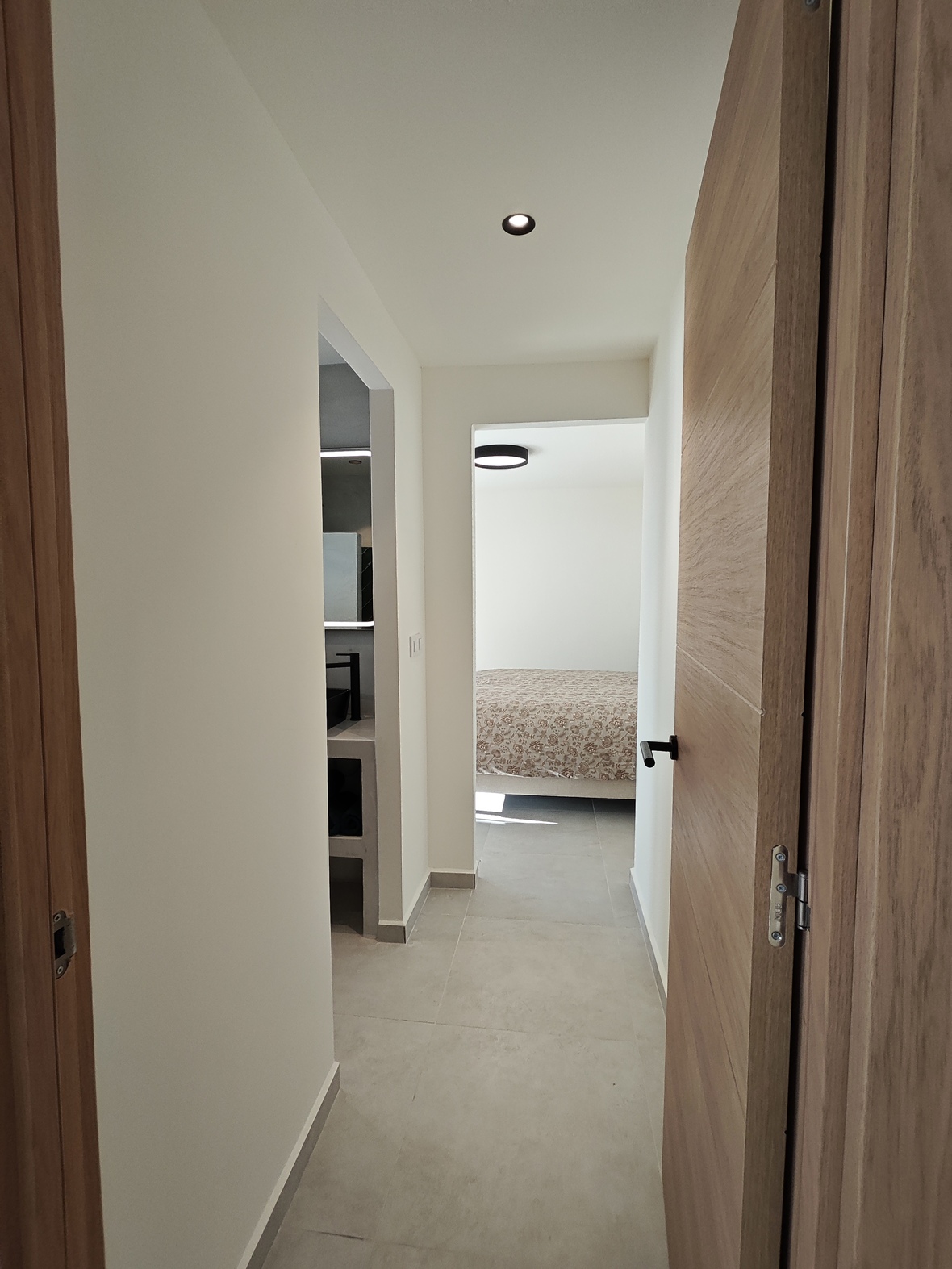 Moderne gang met licht houten deuren en grijze tegelvloer, die toegang biedt tot een minimalistische badkamer en een lichte slaapkamer.