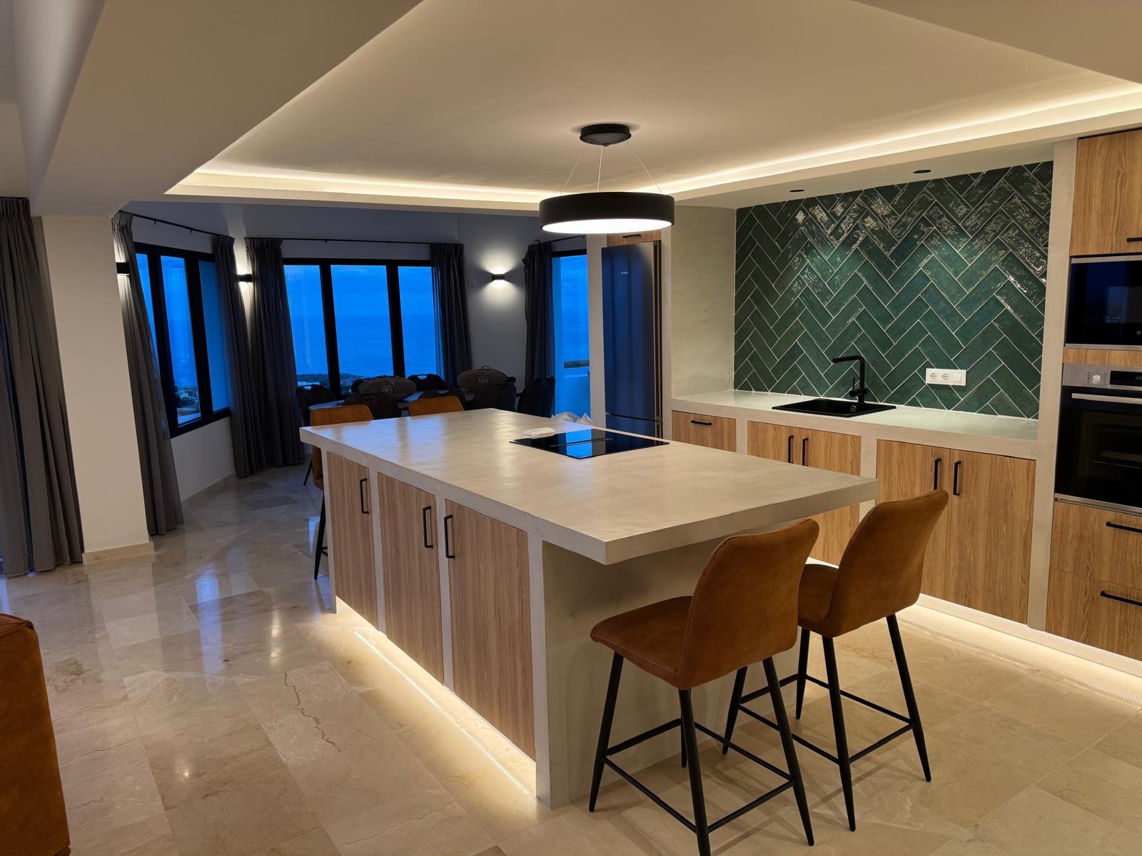 Moderne keuken met een eiland van microcement, groene achterwand in visgraatmotief en panoramisch uitzicht op zee door grote ramen tijdens de schemering.