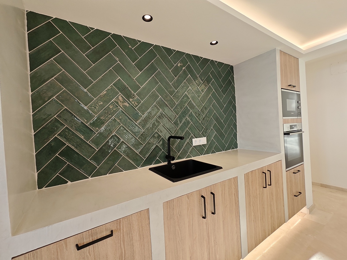 Moderne keuken met een groene visgraat tegelwand, zwarte spoelbak en licht houten kasten met inbouwapparatuur en indirecte verlichting.