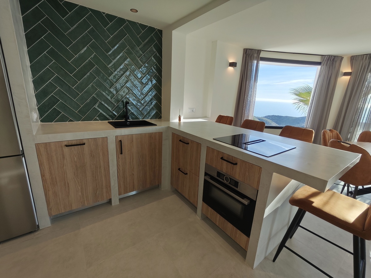 Moderne keuken met groene visgraat achterwand, houten kasten en een kookeiland met geïntegreerde kookplaat en panoramisch uitzicht op zee.