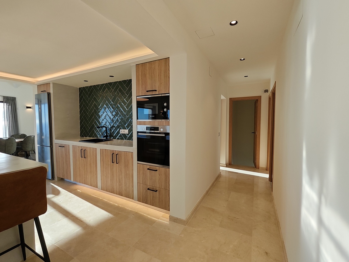 Moderne keuken met houten kasten, donkergroene achterwand in visgraatmotief en inbouwapparatuur, grenzend aan een lichte gang met natuurstenen vloer.