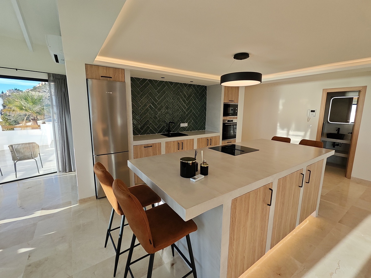 Moderne keuken met kookeiland, stenen werkblad, lederen krukken en een donkergroene achterwand in visgraatmotief. Voorzien van inbouwapparatuur en toegang tot het terras.