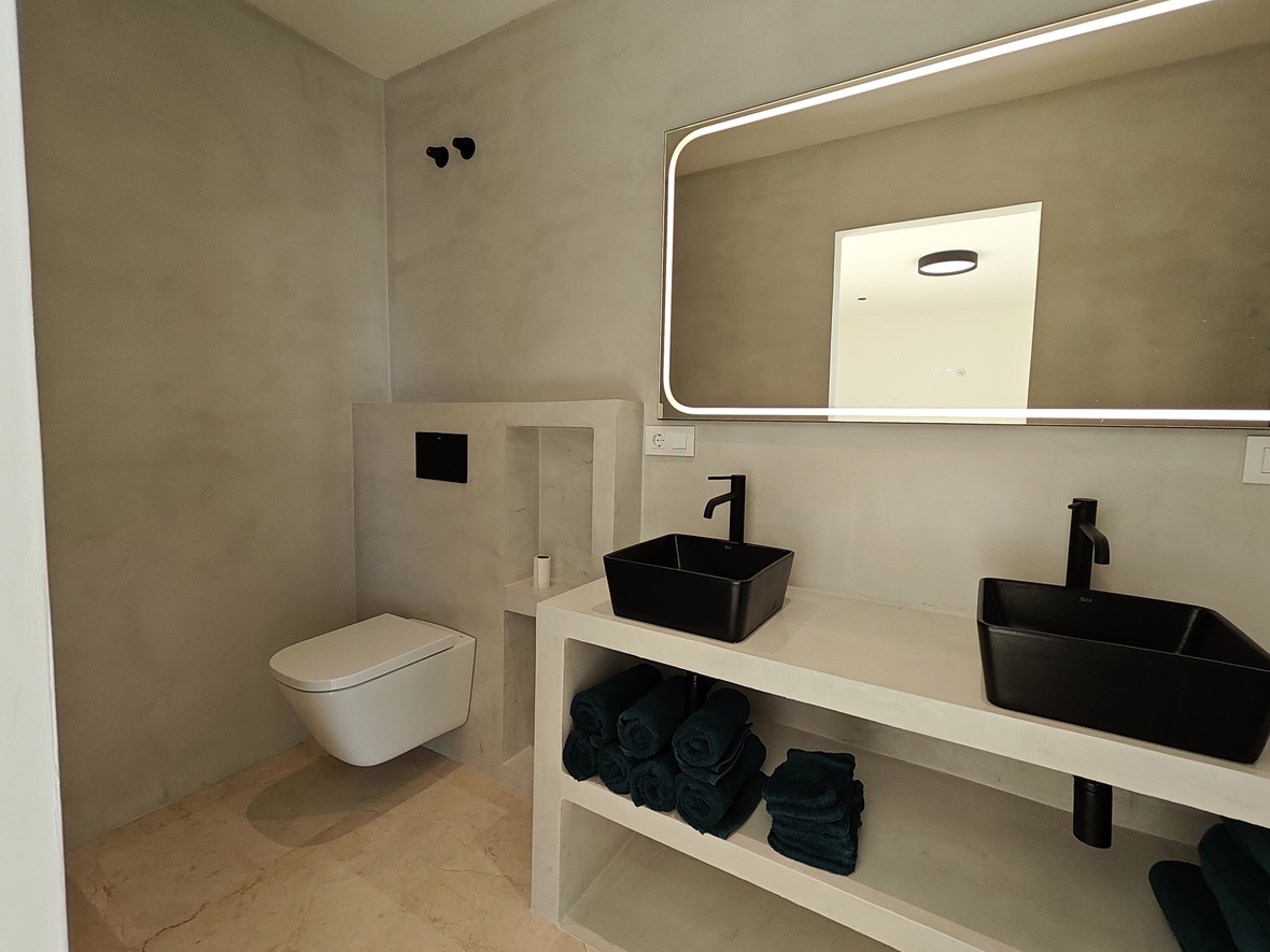 Moderne minimalistische badkamer met grijze wanden van microcement, dubbele zwarte opzetwastafels en een grote spiegel met geïntegreerde LED-verlichting.