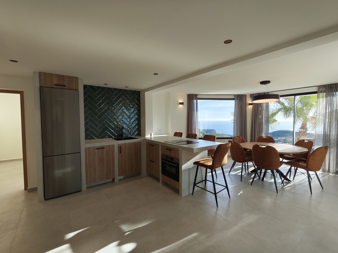 Moderne open keuken met groene visgraat achterwand, inbouwapparatuur en een eethoek met panoramisch uitzicht op zee door grote raampartijen.
