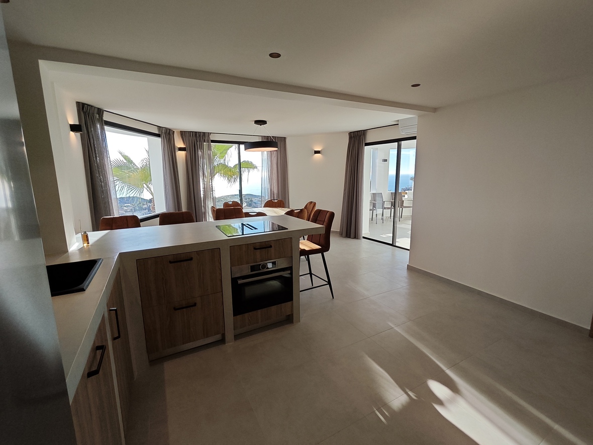 Moderne open keuken met houten kasten, kookeiland en eethoek met panoramisch uitzicht op zee via grote raampartijen en schuifdeuren naar het terras.