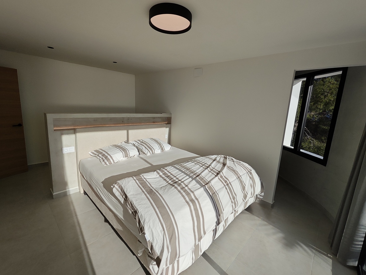 Moderne slaapkamer met een op maat gemaakt hoofdbordpaneel inclusief LED-verlichting, grootformaat vloertegels en een raam met zwart profiel en uitzicht op bomen.