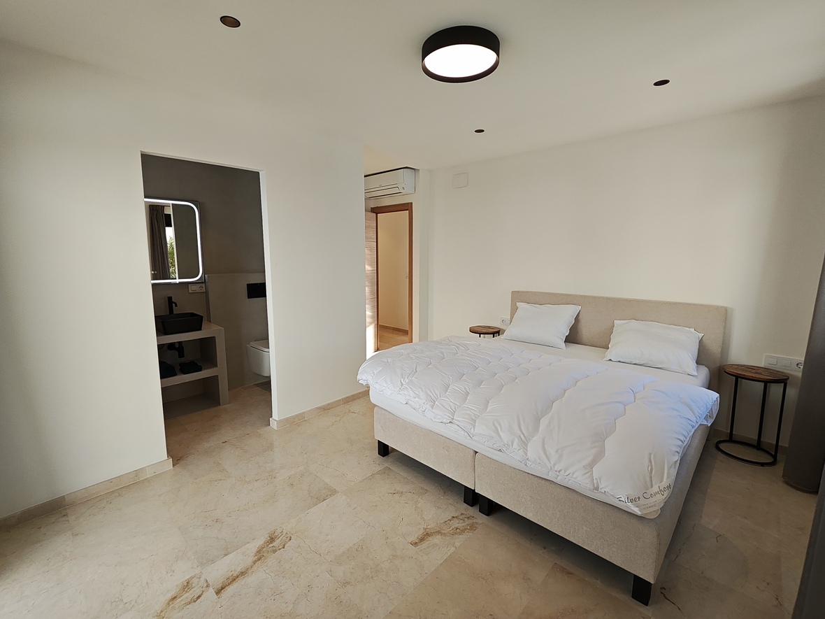 Moderne slaapkamer met marmeren vloer, gestoffeerd tweepersoonsbed en een en-suite badkamer met zwarte opzetwastafel en verlichte spiegel.
