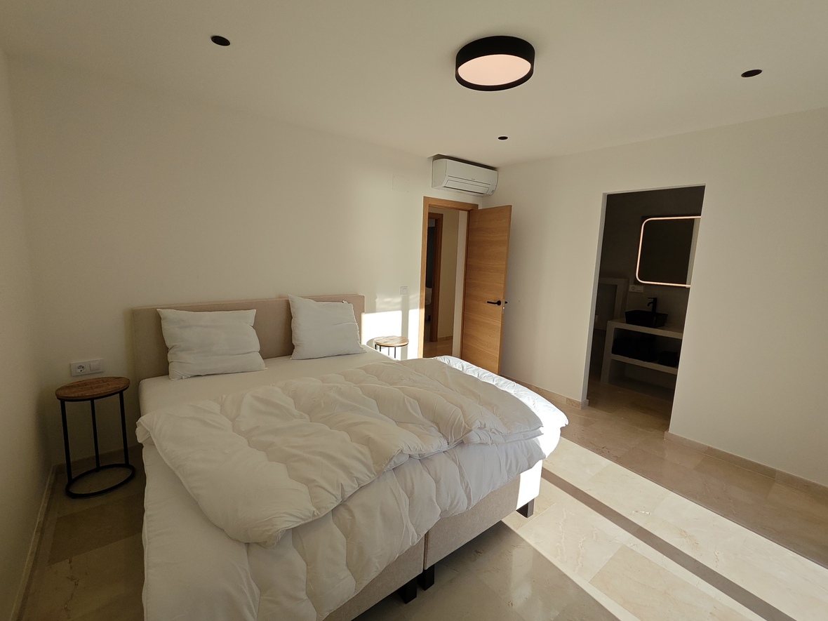 Moderne slaapkamer met stenen tegelvloer, beige gestoffeerd hoofdbord en minimalistische bijzettafels. Voorzien van airconditioning en een en-suite badkamer met verlichte spiegel.
