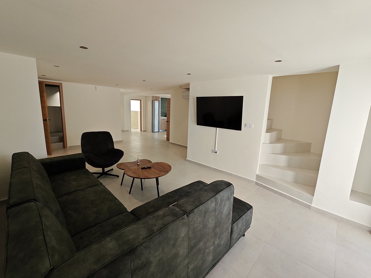 Moderne woonkamer met donkere hoekbank, houten salontafel en wand-tv, grenzend aan een minimalistische witte trap met inbouwspots.