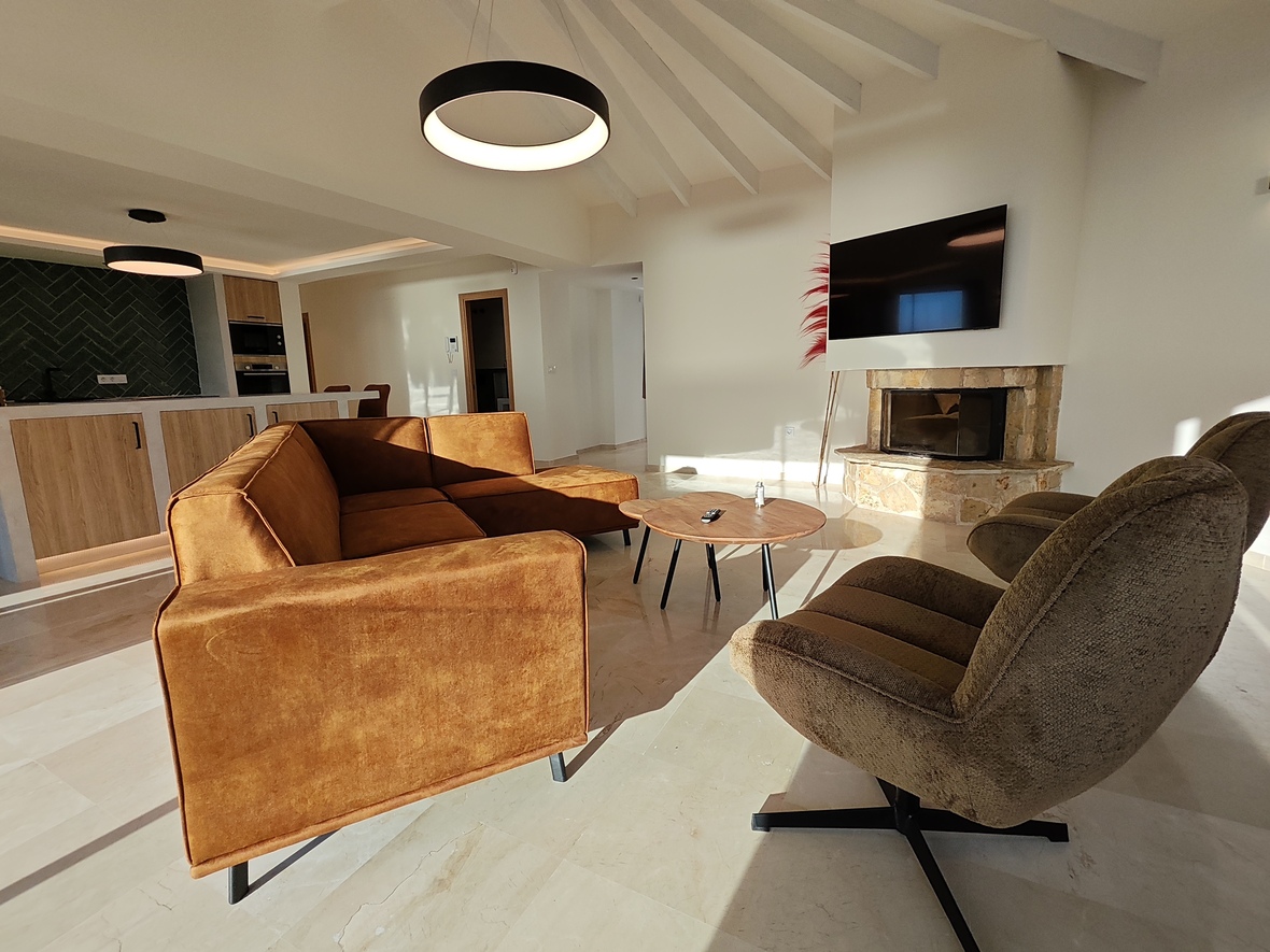 Moderne woonkamer met okerkleurige fluwelen bank, natuurstenen haard en wit balkenplafond. Op de achtergrond de open keuken met groene visgraat tegels en gepolijste stenen vloeren.