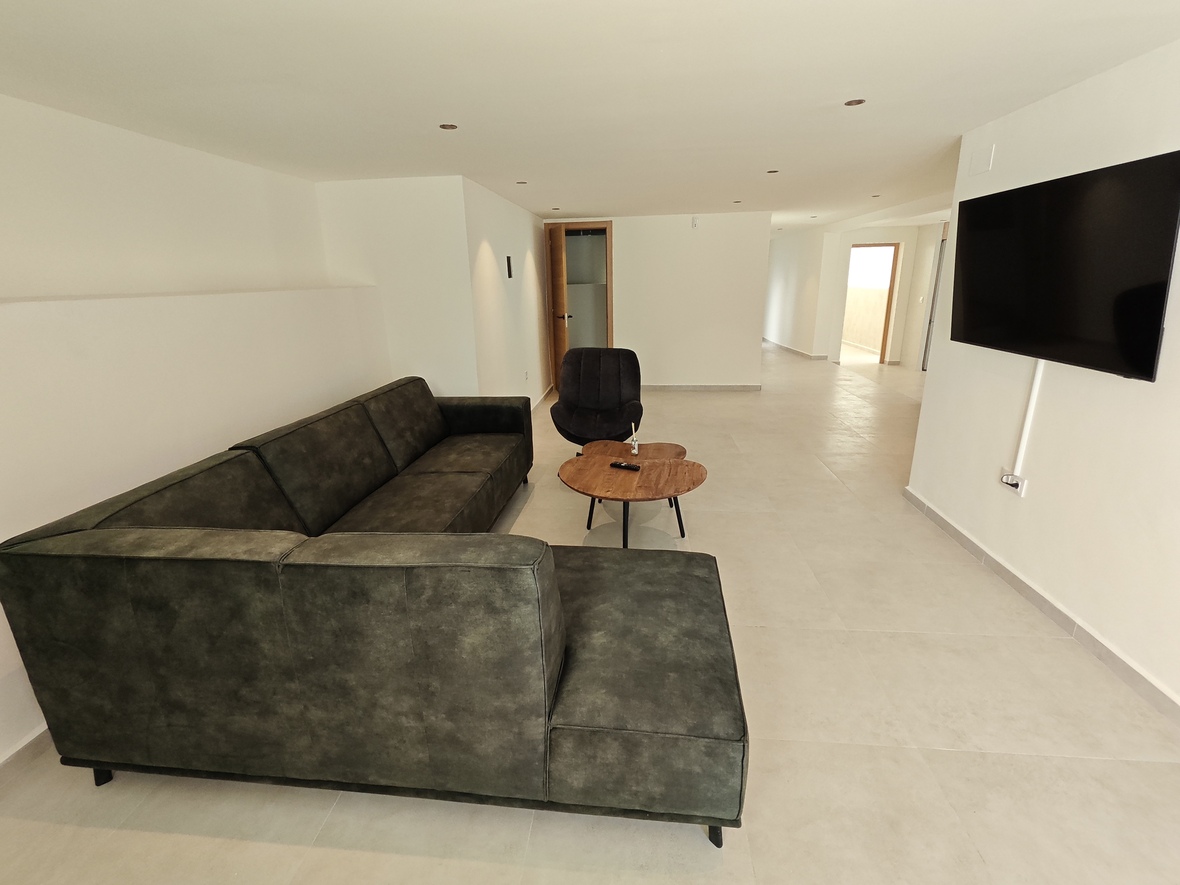 Moderne woonruimte met een donkergroene hoekbank, natuurhouten salontafel en aan de wand gemonteerde flatscreen-tv op een lichte tegelvloer.