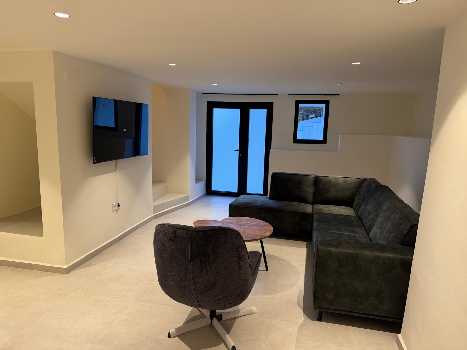 Moderne zithoek met donkere hoekbank, draaifauteuil en wandtelevisie. Inbouwspots en een neutrale tegelvloer accentueren de strakke, minimalistische afwerking.