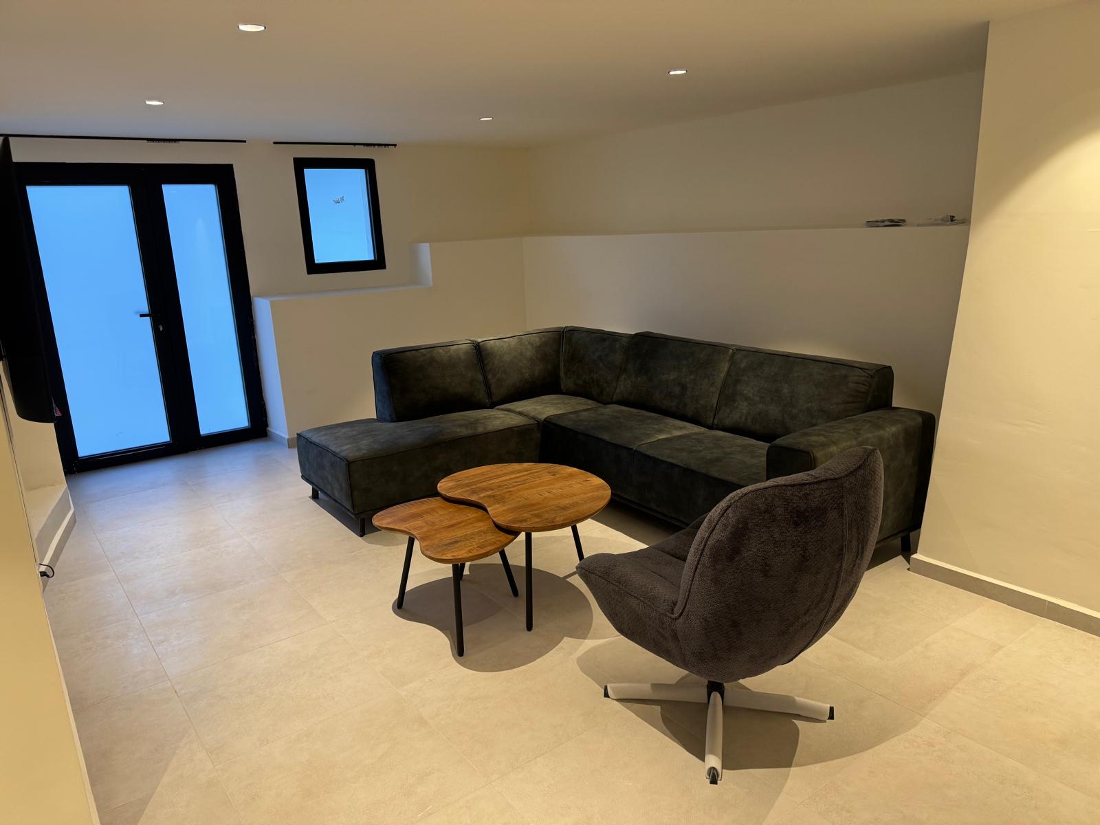 Moderne zithoek met een donkergrijze hoekbank, organische houten salontafels en een draaifauteuil op een lichte tegelvloer met subtiele inbouwspots.