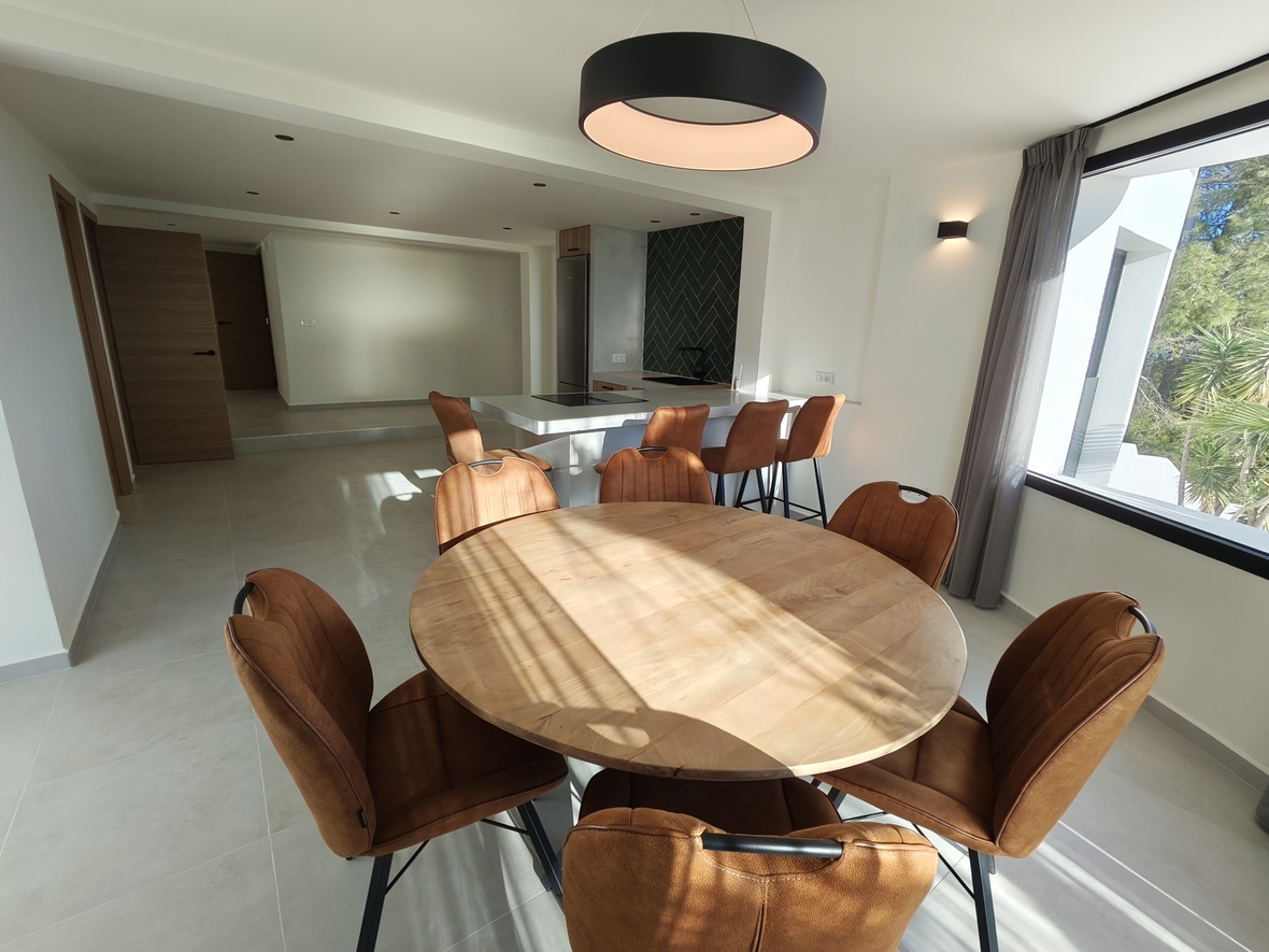 Open eetkamer met een ronde houten tafel, cognacbruine stoelen en een moderne ringlamp, grenzend aan een keuken met kookeiland en groene visgraatwand.