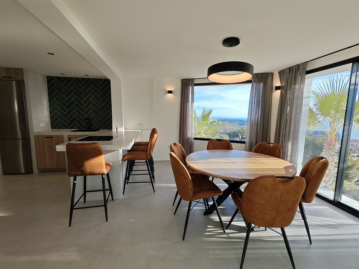 Open eetkamer met ronde houten tafel, cognac stoelen en een moderne keuken met visgraat achterwand en panoramisch uitzicht op zee.