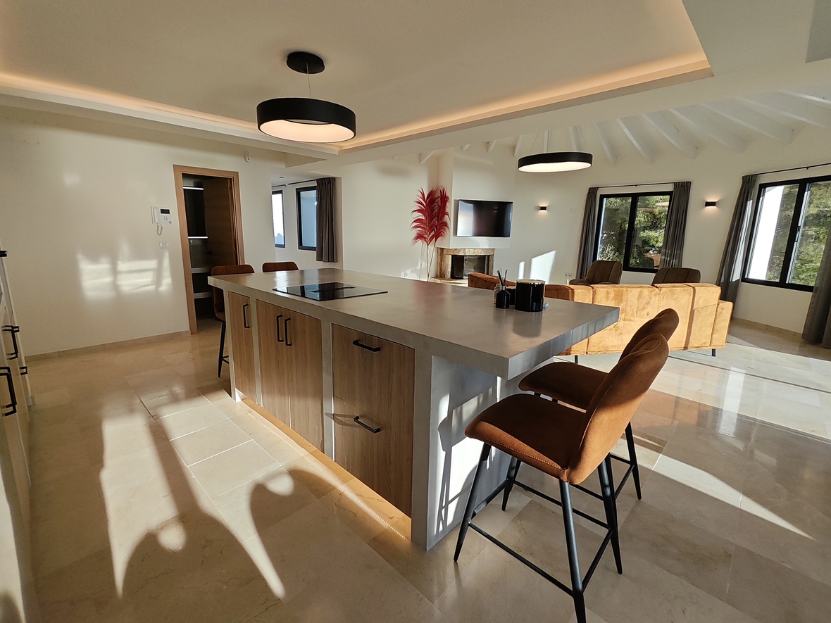 Open keuken met kookeiland in betonlook, inductiekookplaat en lederen barkrukken onder een wit balkenplafond met moderne ronde hanglampen.